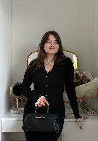 Jeune femme en robe noire tenant un sac à main noir, debout près d'une commode blanche avec des fleurs et un miroir rond.