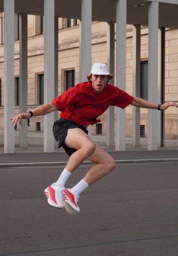 Young man wearing a red shirt, black shorts, and white cap jumps with arms outstretched on a city street with tall pillars behind him.