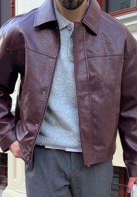 Bărbat îmbrăcat într-o jachetă de piele bordo peste un pulover gri deschis cu nasturi și pantaloni gri, stând în exterior lângă o clădire.