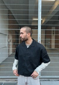 Schwarzes kurzärmeliges Hemd mit Kragen und Brusttasche, über einem hell gestreiften Langarmoberteil getragen. Dazu helle Jeans und Ringe.