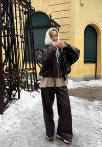 Femme ajustant une capuche en maille blanche, portant une veste marron foncé oversize et un pantalon large en cuir, debout dans une rue enneigée près d'une grille en fer forgé.