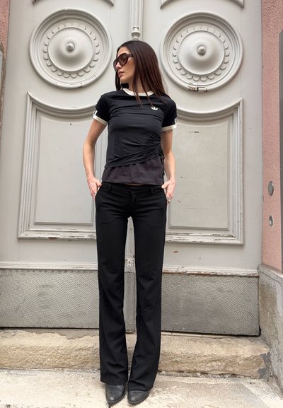Mujer con cabello largo y oscuro que lleva gafas de sol, camiseta negra de Adidas, pantalones negros y botas, de pie frente a una puerta blanca decorativa con detalles circulares.