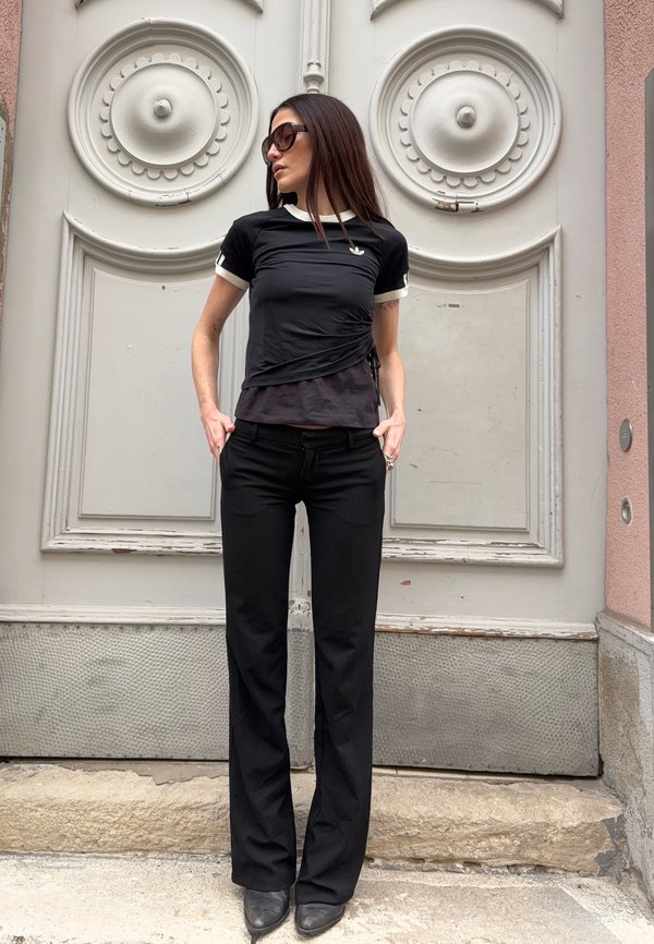 Žena s dlhými tmavými vlasmi, ktorá má na sebe slnečné okuliare, čierne tričko Adidas, čierne nohavice a čižmy, stojí opretá o ozdobné biele dvere s kruhovými detailmi.