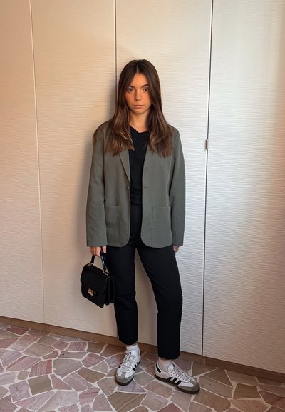 Chaqueta verde oliva con dos bolsillos frontales, blusa negra, pantalones negros y zapatillas grises, complementada con un pequeño bolso negro.