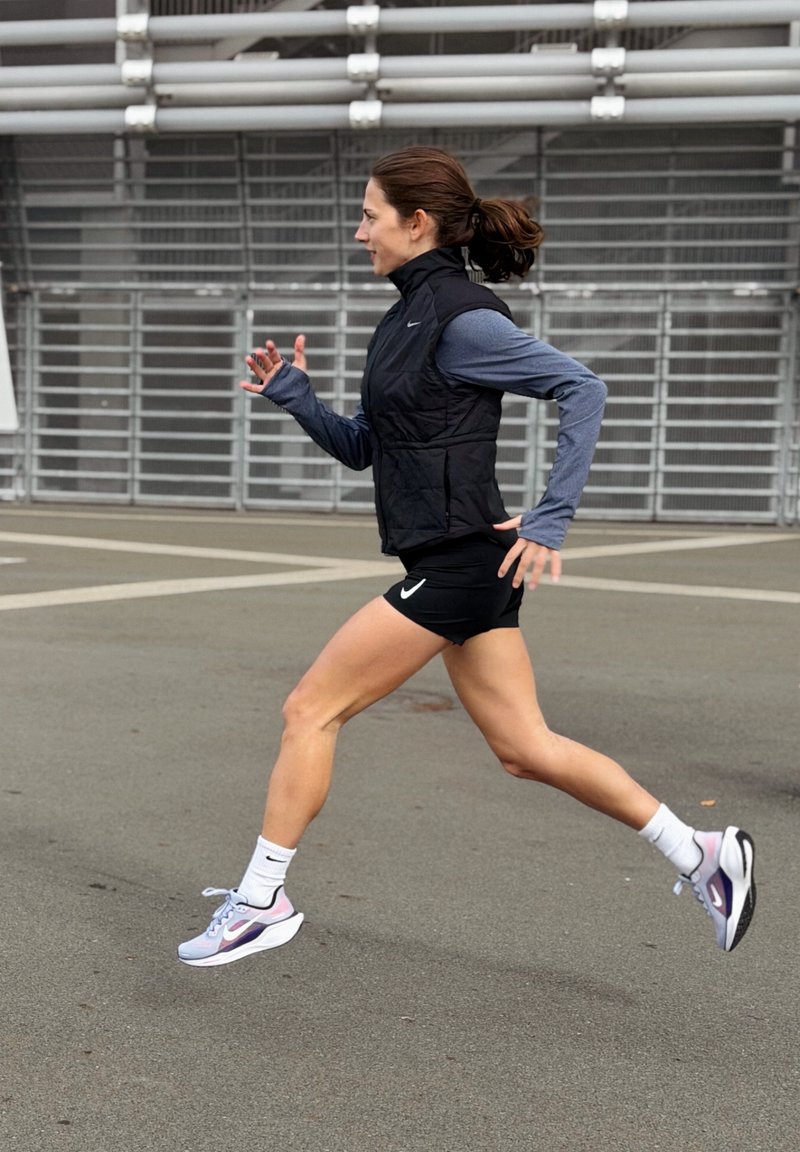 Gilet sans manches noir, haut à manches longues bleu, shorts noirs et chaussures de course accentuées de violet. L'arrière-plan comprend du pavé gris et des structures métalliques.