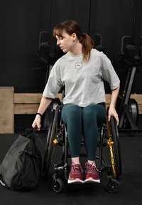 Een persoon in een rolstoel draagt een grijs t-shirt en teal groene legging en reikt naar een zwarte tas. De rolstoel heeft gouden accenten en zwarte wielen.
