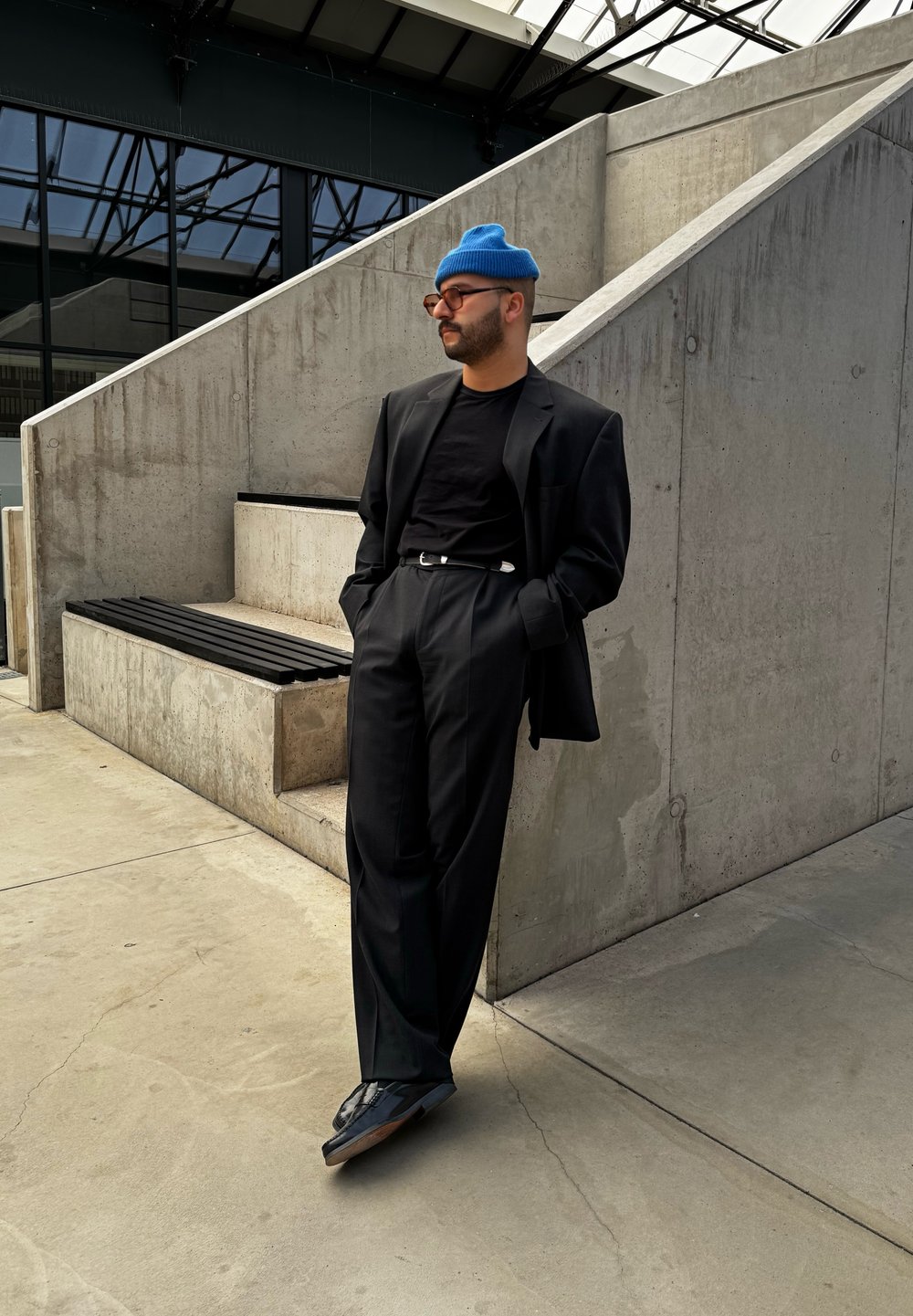 Μαύρο σακάκι πάνω από μαύρο μπλουζάκι, συνδυασμένο με ψηλόμεσες μαύρες παντελόνες. Ένα μπλε σκουφί και μαύρα παπούτσια ολοκληρώνουν την εμφάνιση. Φόντο από σκυρόδεμα.