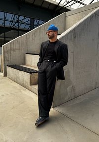 Μαύρο σακάκι πάνω από μαύρο μπλουζάκι, συνδυασμένο με ψηλόμεσες μαύρες παντελόνες. Ένα μπλε σκουφί και μαύρα παπούτσια ολοκληρώνουν την εμφάνιση. Φόντο από σκυρόδεμα.