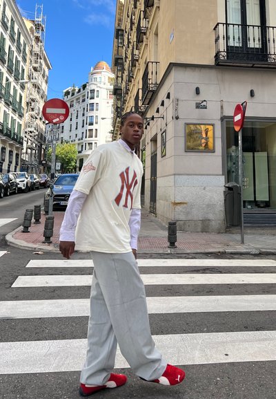Camiseta blanca con el logo de NY, pantalones de pierna ancha en gris claro y zapatillas rojas. Entorno urbano con edificios y señales de tráfico en el fondo.