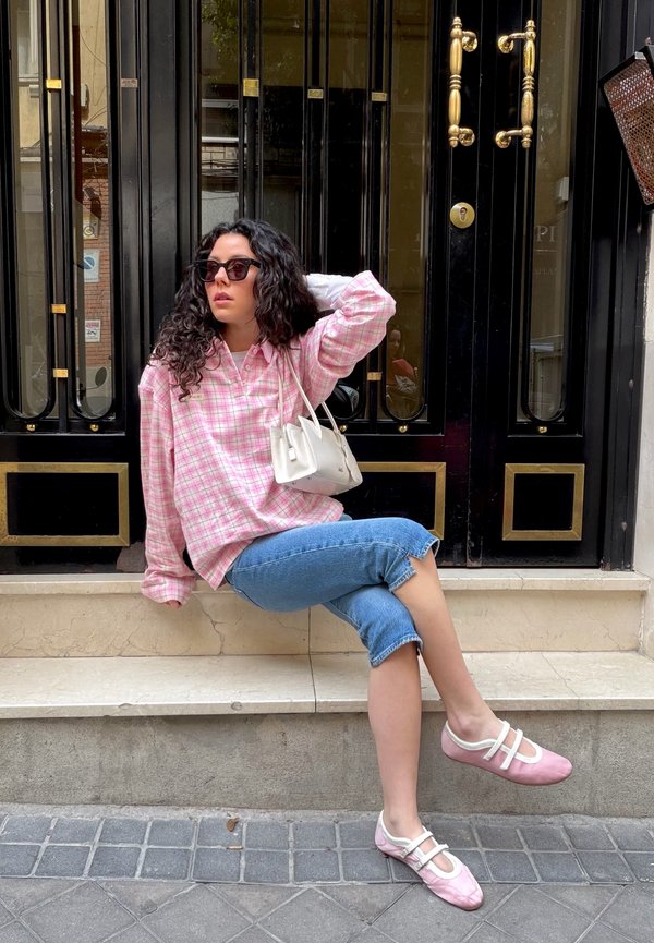 Femme aux cheveux bouclés portant des lunettes de soleil, une chemise à carreaux rose, un jean court et des chaussures roses, assise sur les marches devant une porte noire et dorée.