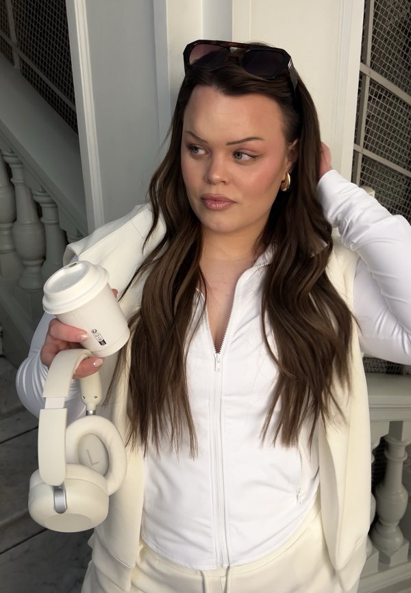 Femme aux longs cheveux bruns portant une veste blanche zippée, tenant une tasse de café et des écouteurs blancs, s'ajustant les cheveux, portant des lunettes de soleil sur la tête.