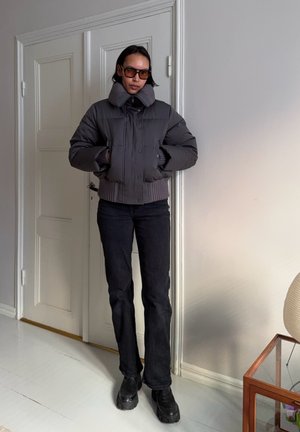 Persona de pie en el interior vistiendo una chaqueta acolchada oscura, pantalones negros, botas negras gruesas y gafas de sol marrones, con las manos en los bolsillos.