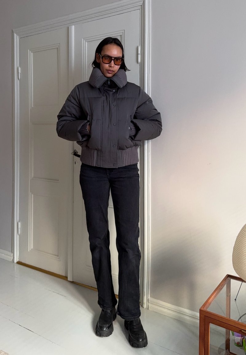 Personne debout à l'intérieur portant une veste matelassée foncée, un pantalon noir, des bottes noires massives et des lunettes de soleil marron, les mains dans les poches.