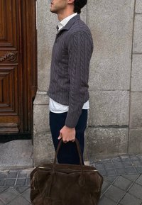 Homme en pull gris à torsades et chemise blanche tenant un grand sac en suède marron, debout à côté d'un mur en pierre et d'une porte en bois.