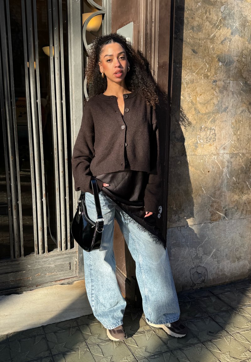 Femme aux cheveux bouclés portant un cardigan marron foncé, un haut noir, un jean large bleu clair et des baskets, tenant un sac à main noir en plein air.