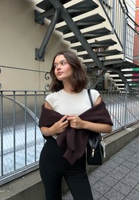 Világos bézs, testhezálló póló, sötétbarna pulóver a karokon, fekete, magas derekú leggings, fekete táska gyöngyökkel díszítve, városi környezet.