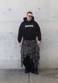 Svart hettegenser med hvit grafisk tekst, lagdelt rutete skjørt i gråblå toner, svarte vide bukser, speilvendte solbriller og synlige tatoveringer.