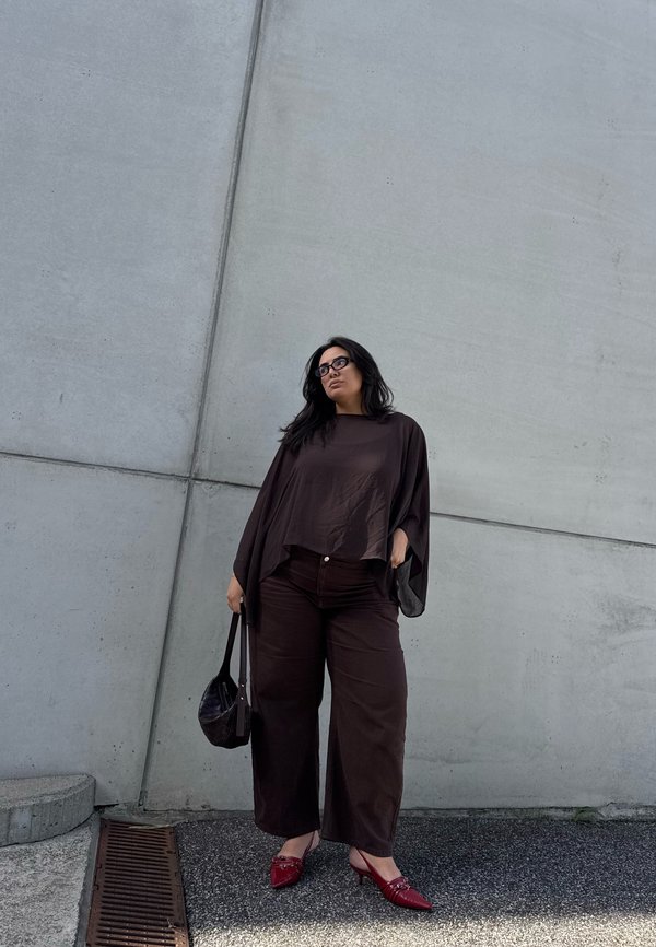 Mujer con cabello largo y negro que lleva pantalones anchos y oscuros y una blusa, tacones rojos puntiagudos y gafas, está de pie junto a una pared de hormigón sosteniendo una bolsa negra.
