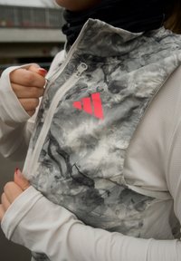 Gilet sans manches gris et blanc avec un motif marbré, comportant un logo rose et une fermeture éclair. La couche de base présente un design texturé blanc.