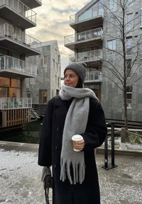Casaco preto, cachecol cinza com franjas, gorro cinza de malha. Segurando um copo de papel de café. Edifícios modernos e chão coberto de neve ao fundo.