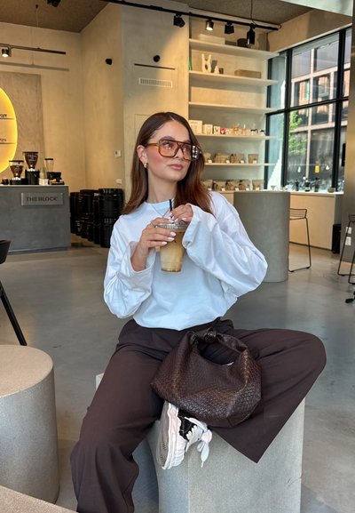 Modelo que lleva una camisa blanca de manga larga y pantalones marrones, sosteniendo una bebida fría. Los accesorios notables incluyen gafas de gran tamaño y un bolso de mimbre marrón.