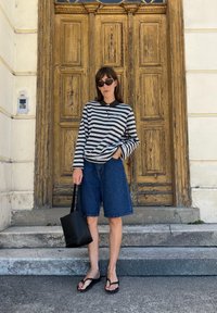 Mulher vestindo uma camisa de mangas longas listrada, calções de ganga, chinelos e óculos de sol, está a segurar uma bolsa preta diante de grandes portas de madeira.