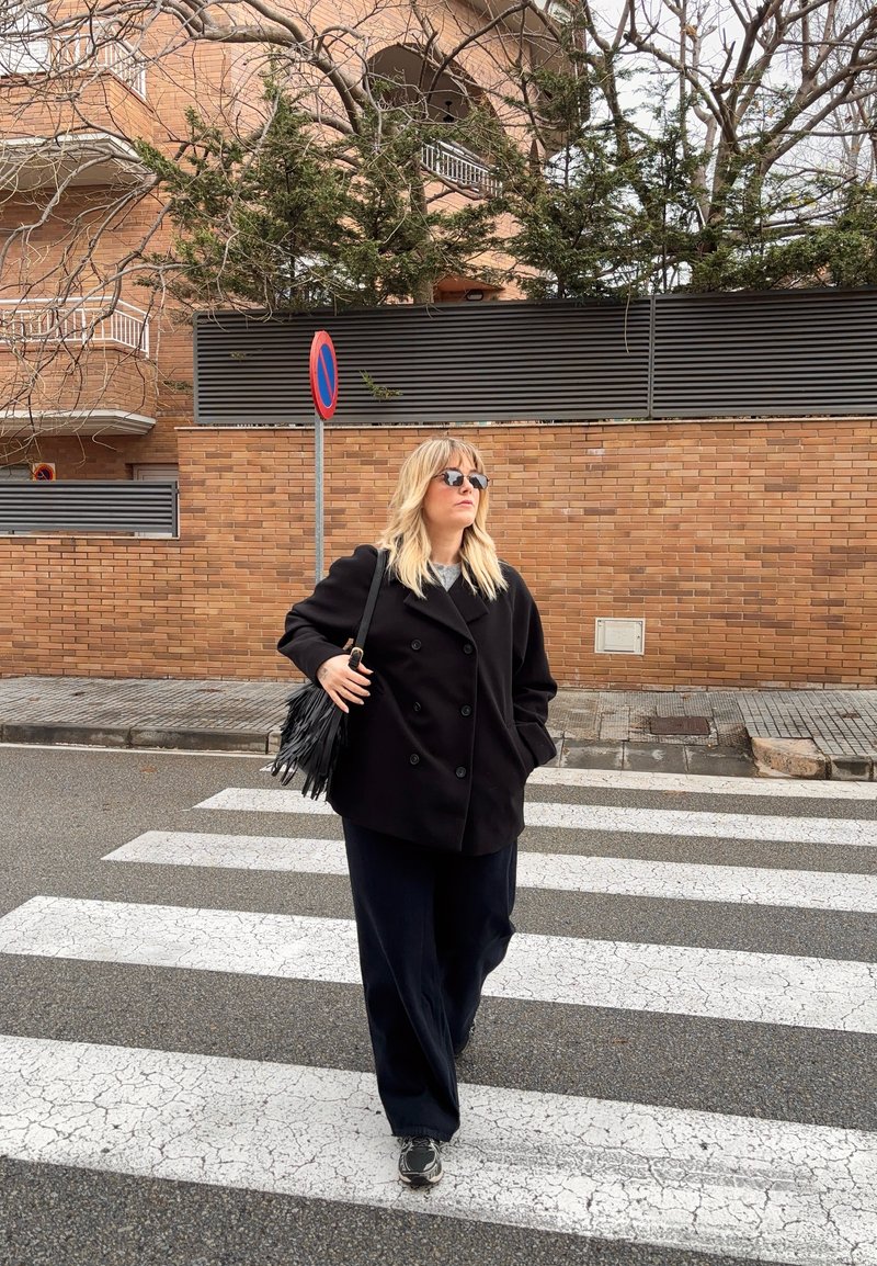 Femme portant un manteau noir et des lunettes de soleil marchant sur un passage piéton près d'un bâtiment en briques et d'un panneau d'interdiction de stationnement.
