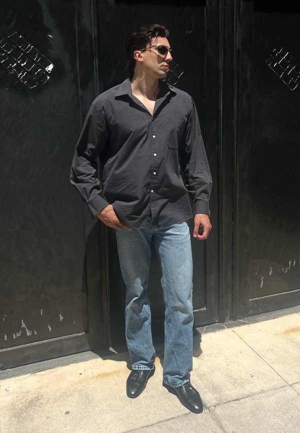 Mann trägt schwarzes, geknöpftes Hemd, blaue Jeans, schwarze Schuhe und Sonnenbrille und steht vor einer dunklen Metalltür auf einem sonnigen Gehweg.