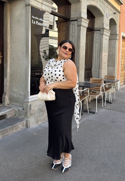 Falda de satén negra combinada con una blusa blanca de lunares y un pañuelo. Sostiene un bolso de mano crema, lleva zapatos de encaje en blanco y negro y gafas de sol.