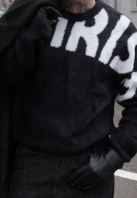 Personne portant un pull noir duveteux avec de grandes lettres blanches, des gants en cuir noir, et tenant un manteau noir sur l'épaule.