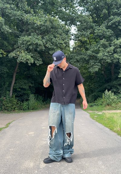 Camisa negra de manga corta con un patrón texturizado, combinada con jeans holgados de un azul claro desgastados que presentan grandes desgastes. Zapatillas negras y gorra.