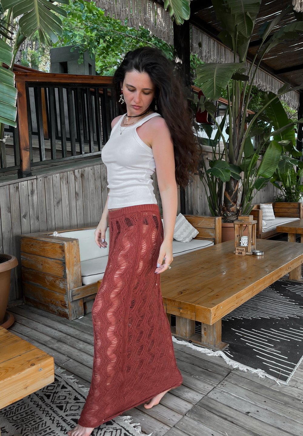 Duga, rust-boja čipkana maksi suknja sa valovitim uzorcima, uparena sa uskim, bijelim, bezrukavim topom. Prirodni drveni namještaj u pozadini.