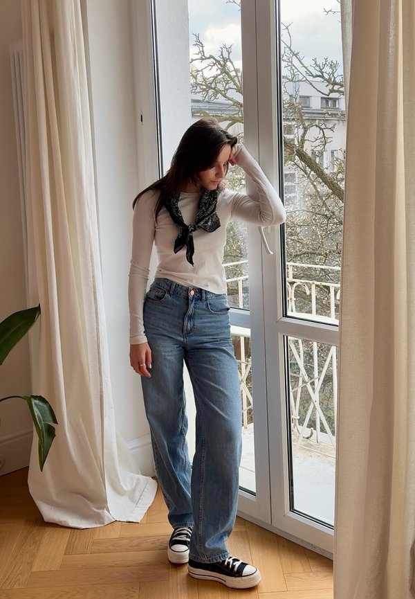 Vrouw in jeans, wit shirt met lange mouwen en een zwarte sjaal staat bij een glazen deur, kijkt naar beneden met één hand op haar hoofd binnen.