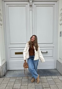 Kvinde i en cremet jakke over en brun sweater og blå jeans står nær hvide dobbeltdøre. Hun holder en brun håndtaske. Gulvet er belagt med fliser.