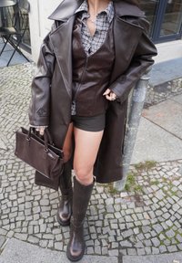 Trench coat maro din piele peste un vestă mulată din piele, asortat cu pantaloni scurți închiși la culoare și cizme maro până la genunchi. Poartă o geantă mică maro.