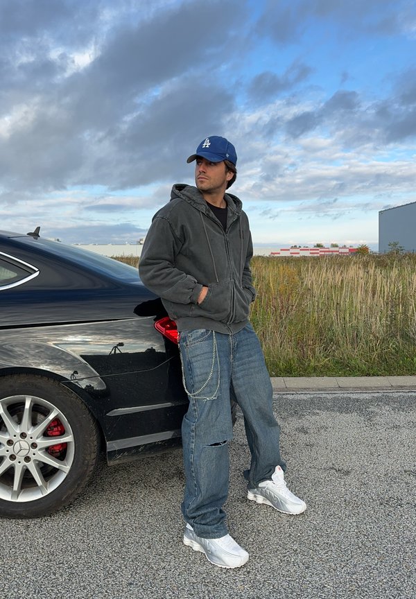 Sivá mikina na zips, čierne tričko, roztrhnuté modré džínsy s retiazkou, biele tenisky. Čierne auto v pozadí, zamračené nebo.
