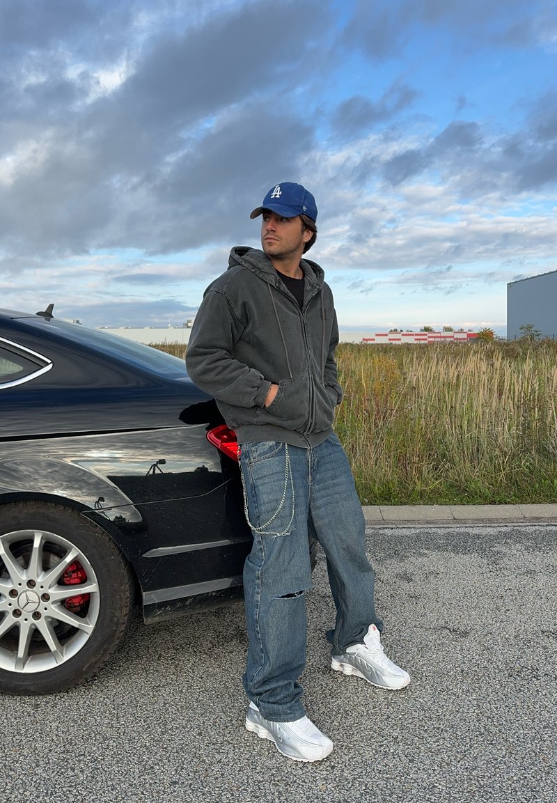 Γκρίζα φούτερ με φερμουάρ, μαύρη μπλούζα, φθαρμένα μπλε τζιν με αλυσίδα, λευκά αθλητικά παπούτσια. Μαύρο αυτοκίνητο στο παρασκήνιο, συννεφιασμένος ουρανός.