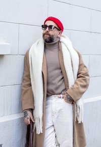 Hnedý vlnený kabát cez béžový rolák, biely šál a bledé nohavice. Červená pletená čiapka a čierne okuliare, s vyznačenými tetovaniami na ruke.