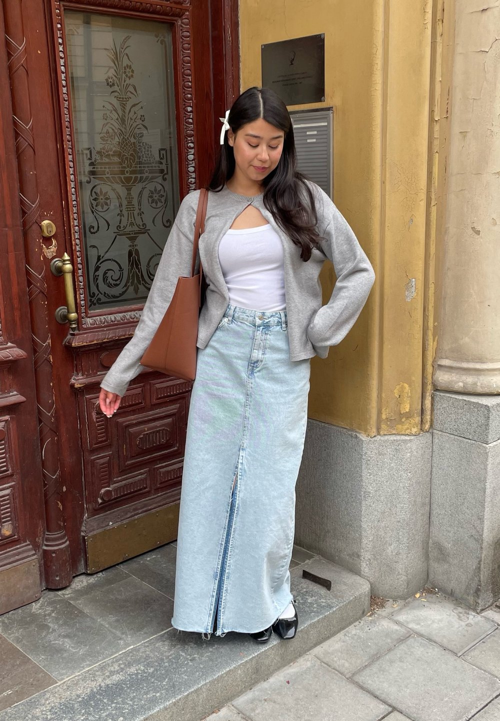 Cardigan cinza crop, top branco, saia maxi em denim azul claro com fenda frontal, sapatos de salto pretos e uma bolsa tote castanha. Ambiente casual ao ar livre.