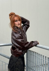 Rjava umetna usnjena jakna s sproščenim krojem in gumbom za zapiranje, v kombinaciji s črno čipkastimi krilom. Model pozira ob metalni ograji.