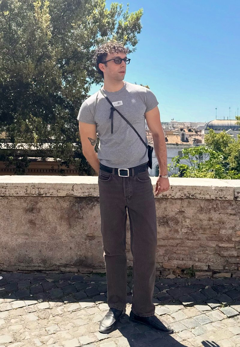 Mand iført briller, grå t-shirt, mørke jeans og sorte sko står på brostensbelagt gade ved stenmuren med bylandskab og træer bagved.