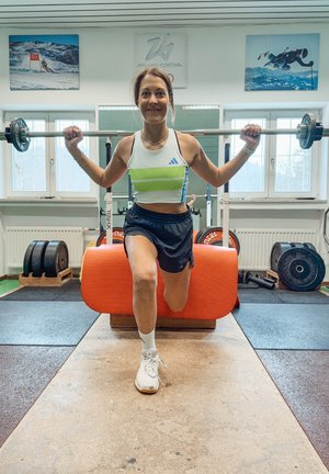 Una persona in palestra esegue un affondo mentre tiene un bilanciere. Indossa un top corto atletico bianco e verde e pantaloni corti neri.