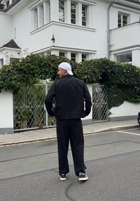 Svart denimjakke, løse svarte bukser og svarte og hvite sneakers. En hvit caps fullfører antrekket. Bakgrunnen viser en hvit bygning med grønn beplantning.