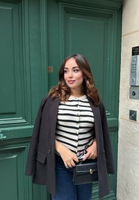 Person in einem schwarzen Blazer über einem cremefarbenen und schwarzen gestreiften Pullover, hält eine kleine schwarze Handtasche. Im Hintergrund ist eine grüne Tür zu sehen.