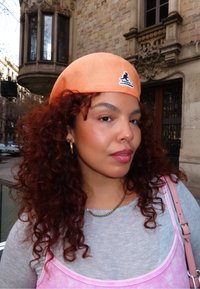 Femme aux cheveux roux bouclés portant une casquette Kangol orange, des boucles d'oreilles en or en forme de créoles, un collier en chaîne dorée, un haut rose par-dessus un t-shirt gris, debout près d'un bâtiment en pierre.