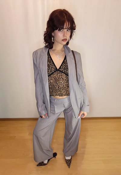 Traje gris de gran tamaño con pantalones de pierna ancha, superpuesto sobre una blusa negra con estampado de leopardo. Los detalles incluyen un collar con colgante en forma de corazón y tacones a rayas.