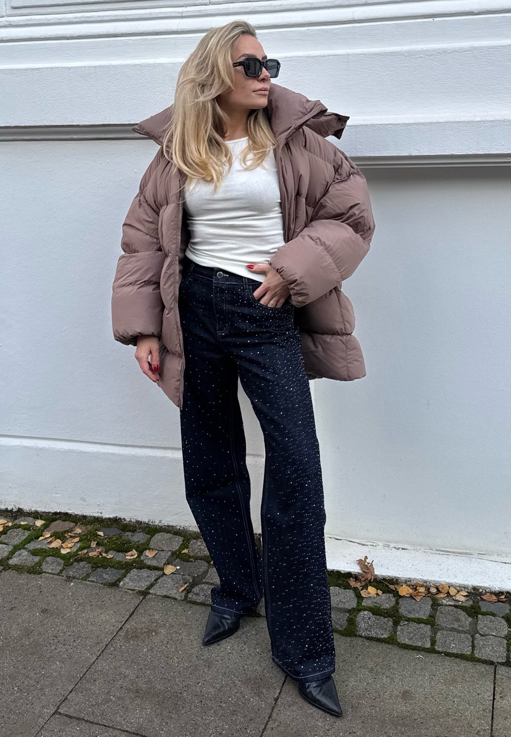 Brun vatteret jakke, hvid top, mørke jeans med pynt og spidse sorte hæle. Modellen poserer mod en hvid væg på en stenmosaiksti.