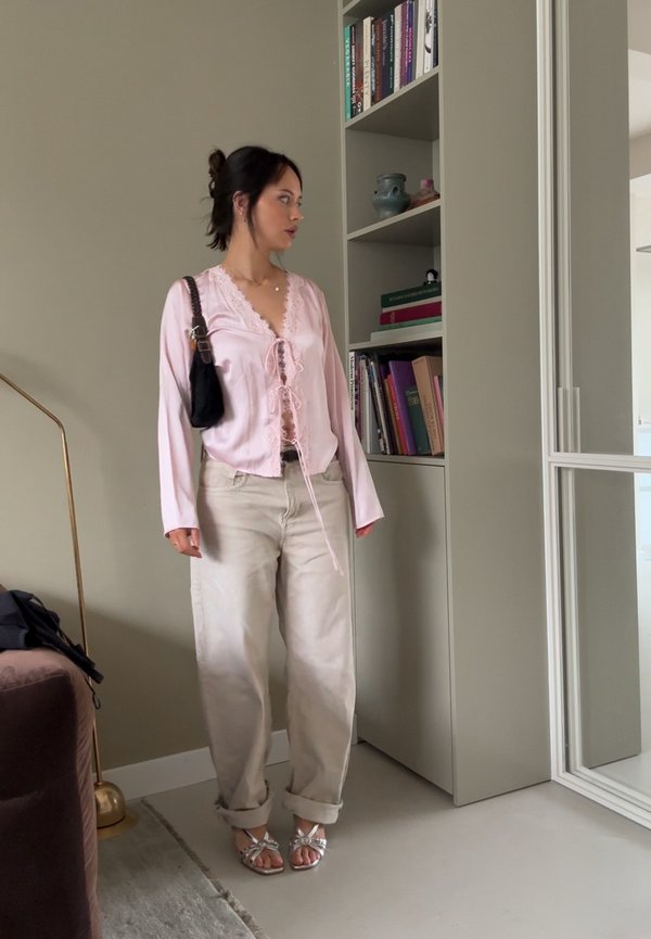 Light pink blouse with lace trim, tied at the front; beige trousers; silver strappy sandals. The background features a bookshelf and a lamp.