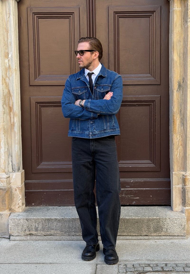 Homme portant des lunettes de soleil, une veste en jean, un pantalon noir et des chaussures noires, debout les bras croisés devant une grande porte en bois marron.