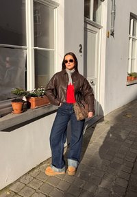 Femme en lunettes de soleil portant une veste en cuir marron, un pull rouge, un jean large et des bottes beige, tenant un sac à imprimé léopard dans une rue pavée ensoleillée.